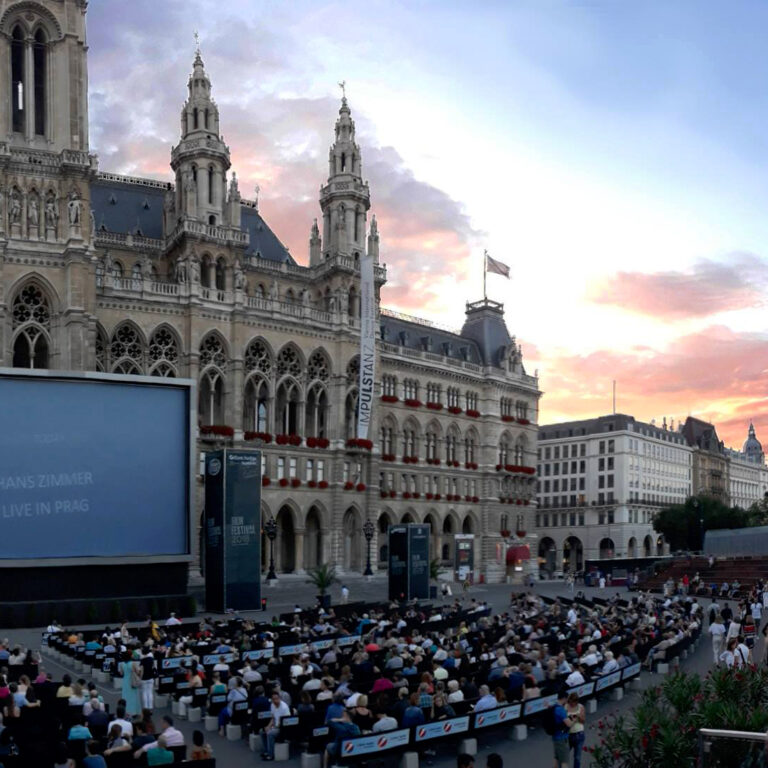 sitzendes Publikum vor einem riesigen Bildschirm vor dem Rathaus bei Sonnenuntergang
